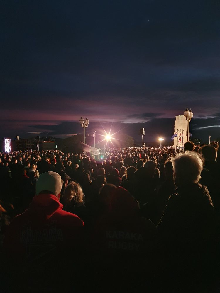 Photograph of a crowd of people gathered in the dark. A stone monument with flags is present to the right, and a bright light shines right at the camera.