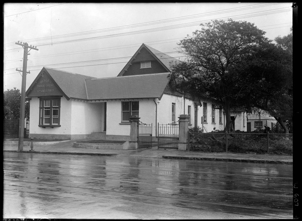 Photograph by Henry Winkelman of the Epsom Public Library in 1924. The image shows what looks like a white stuccoed house with bay window and tiled roof, with the higher gabled roof of a hall rising behind. Auckland Libraries Heritage Collections 1-W0607.
