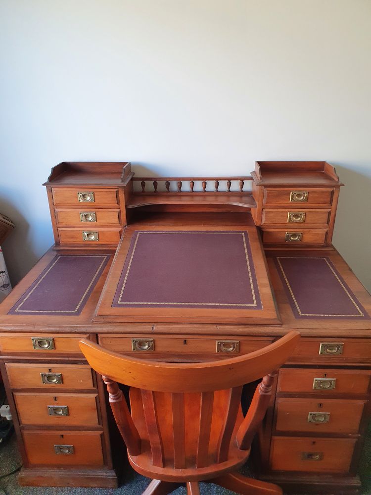 Photograph of a wooden desk with brass handles and three panels of gilt-tooled magenta leather, the middle one of which is a sloped writing surface. At the back corners of the desktop are two sets of three small drawers connected by a galleried shelf. In front of the desk is a slat-backed wooden swivel chair.