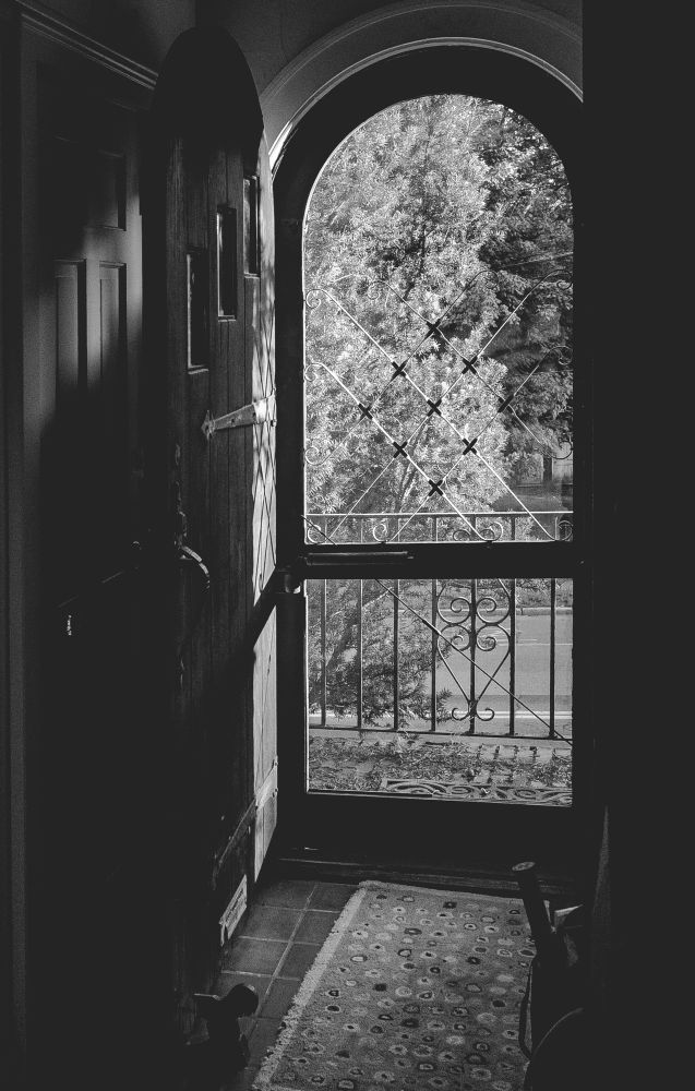 Black and white image of a hallway with an open door, looking out at a sunny day. Not too hot outside. That’s welcome. 