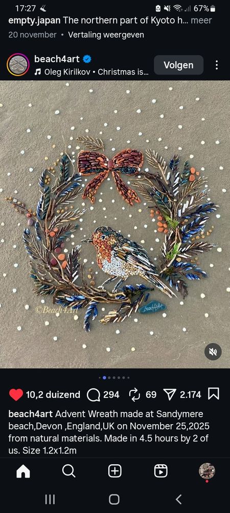 De krans ligt op het zand en is gemaakt van stenen, schelpen en veertjes. Bovenin is een strik te zien. Een roodborstje zit midden in de krans, de pootjes leunen op de onderste rand van de krans.