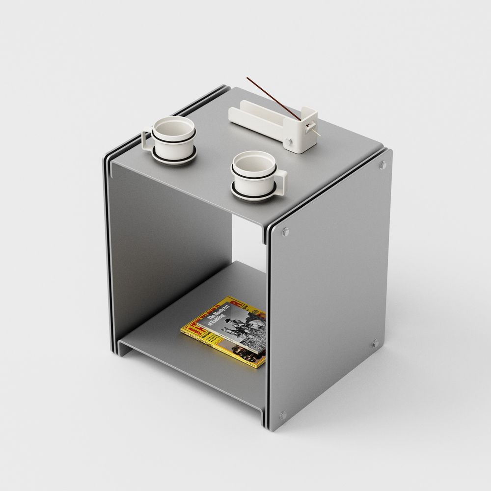 A minimalist gray cubic side table or storage unit with two small white cups on top and a small white incense holder. The cube has an open front with a shelf containing a magazine and a small book. The design features clean lines and a modern aesthetic with visible connection points at the corners.