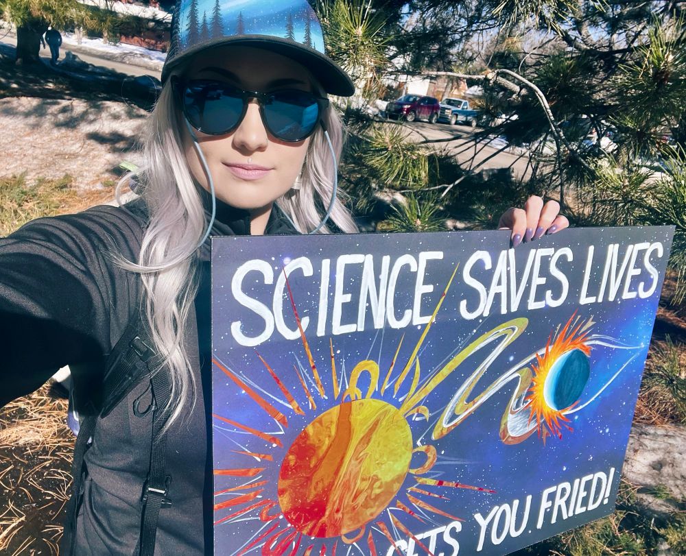 A scientist holding a poster that reads “Science Saves Lives, Denial gets you fried!” with an image of the Sun hitting the Earth with a CME.