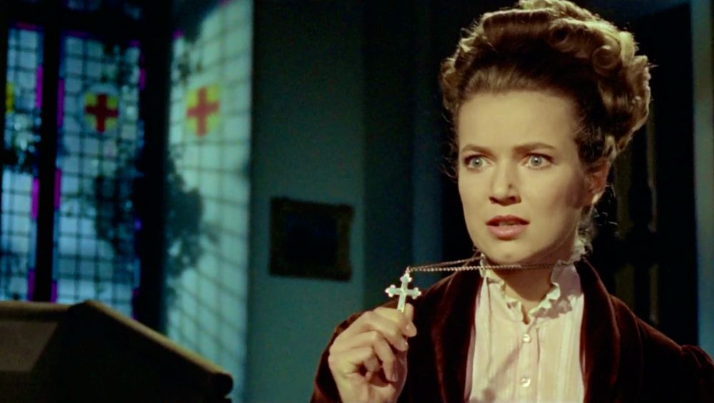 Screenshot of Diana (Suzan Farmer) holding a crucifix up on a chain she’s wearing. She stands aside a stained glass window (also bearing a cross). 