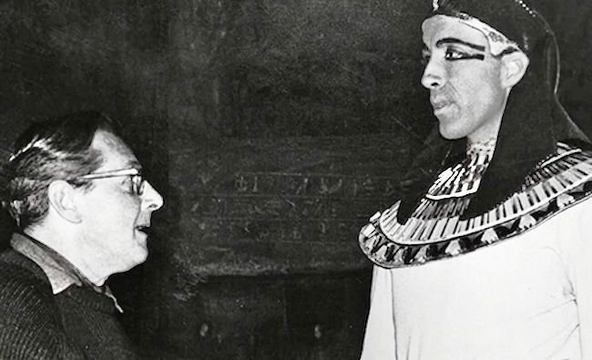 Black and white behind the scenes image. Fisher looks up at/talks to Lee, who’s dressed in an Egyptian headpiece and collar in in black eye make up. 