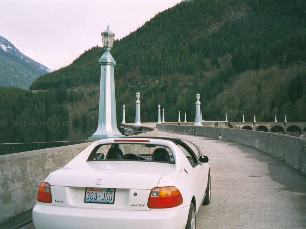 A white 1993 Honda Civic del Sol, a 2-seat targa top with the top removed, viewed from behind atop a curvy dam with old fashioned blue-grey lampposts and evergreen wooded hills in the background on a pleasantly overcast day. 