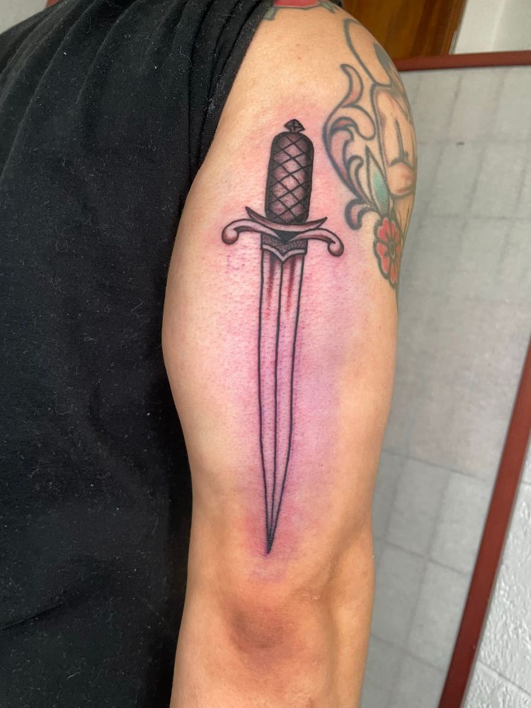 A tattoo of a dagger pointing downwards in black ink with shading on the back of a muscular arm 
