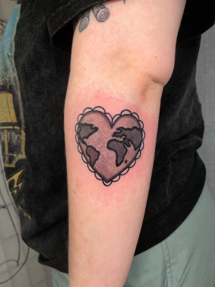 A tattoo on the lower half of the arm, just beneath the elbow and slightly forward, of a heart with the shapes of countries inside resembling a globe. The heart shape has little lace-like half circles surrounding it. The countries are shaded in darker, and the “ocean” part is shaded in with a lighter wash. 
