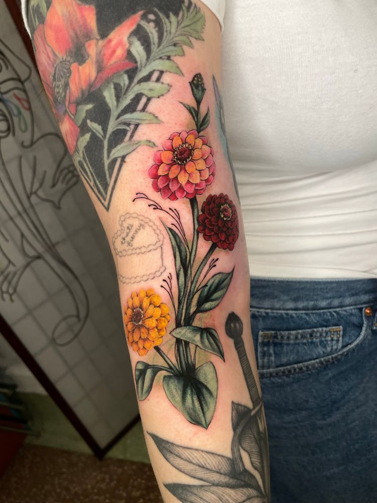 a tattoo on an arm (full of other tattoos) of three zinnias (flowers), the top one in a yellow inside to pink outside gradient, the middle in a deep red and the bottom in yellow. There are leaves coming from the stems and little ornamental plant like sprouts coming out of the main stem area as well. The tattoo is covering up three other tattoos which hopefully you cannot see at all :) 