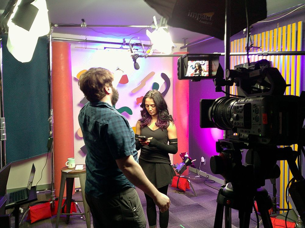 Videographer & Editor Madison adjusts lighting and microphones while Host Niba checks her phone on set. 