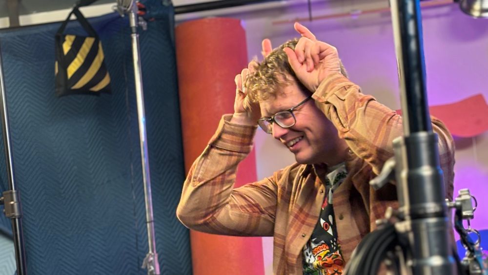 A BTS photo of Hank Green using his forefingers by his temples like bunny ears
