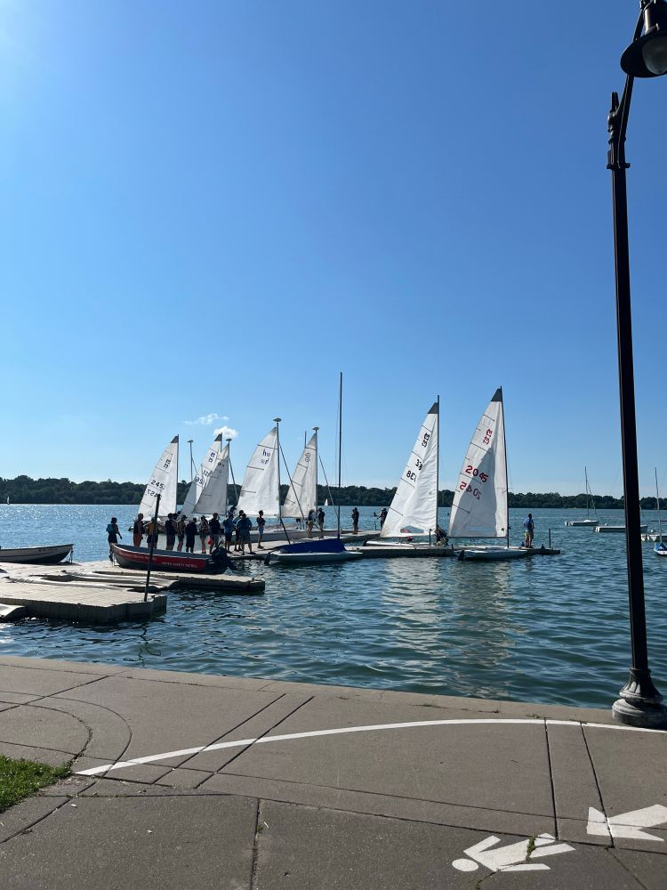 Looking south from the boardwalk around Lake Harriet. In view are 6 sailboats and a variety of brave folks about to board these vessels. 