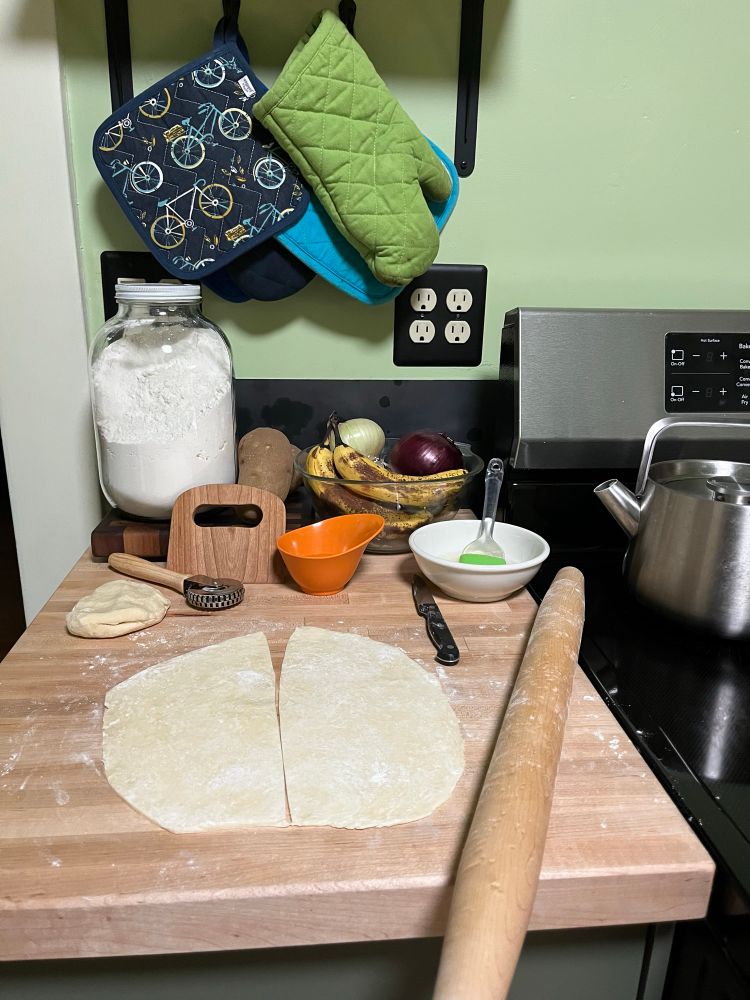 Kitchen counter with rolled out round pie crust cut into two halves. Rolling pin, dough crimper, dough scraper present. 
