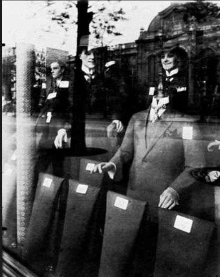 Eugène Atget: Shop Window: Tailor Dummies. This black and white photo from 1910 by Eugene Atget shows a shop window with find suites worn on dummies. But the thing that is really cool is that there is also a reflection of the scene behind this photographer--buildings and the street.