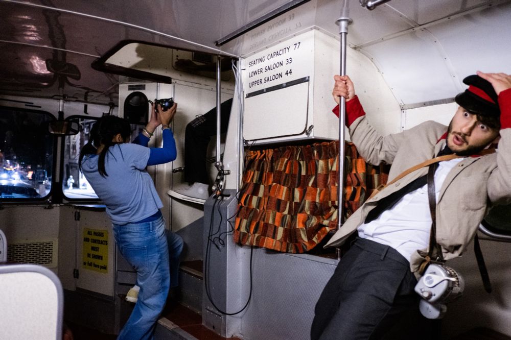 We are on a 1970s bus. A young girl wearing a blue top and blue jeans is clutching a teapot as she tries to steady herself on the bus. A 1970s bus conducter wearing a white shirt and grey trousers and a beige and red conductor jacket has one hand on the bus rail and one hand on his black and red cap as he tries to steady himself on the bus. He has a vintage ticker machine across his body. 