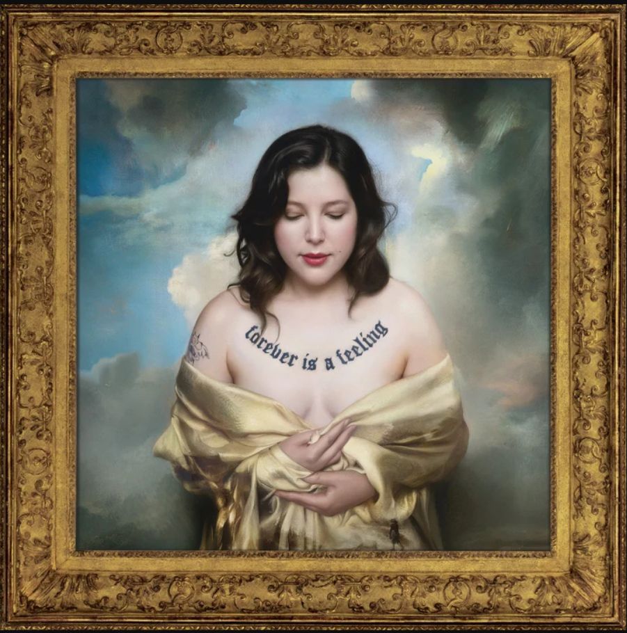 Gorgeous brunette woman in a silky yellow off the shoulder robe with a tattoo saying "forever is a feeling" across her chest standing in front of a cloudy background with a golden frame