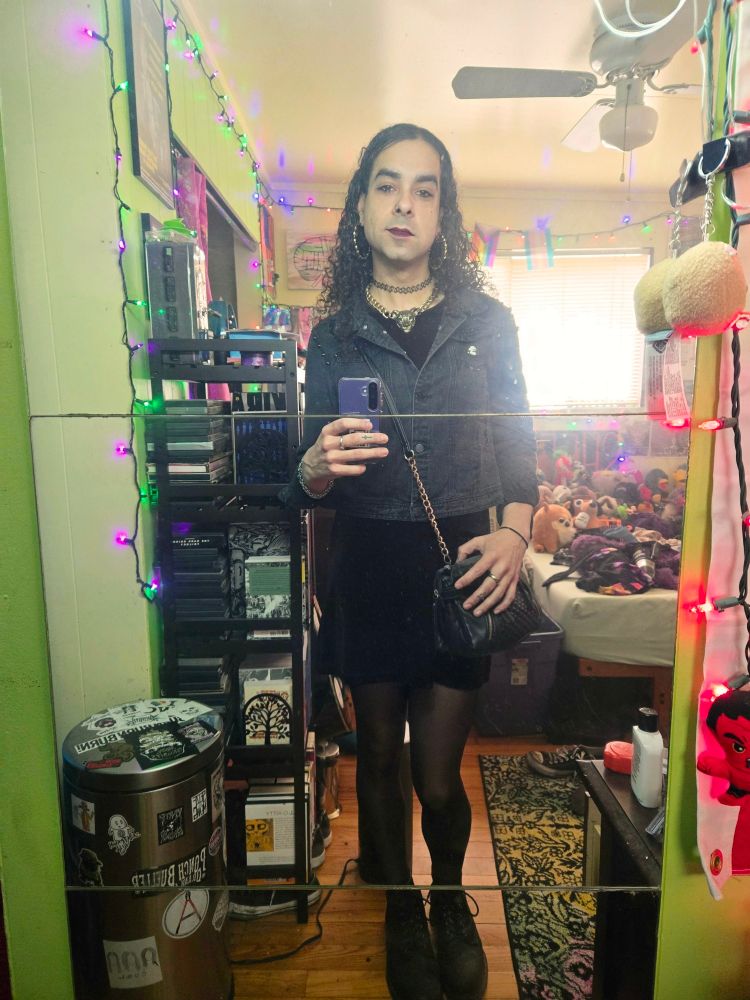 Selfie of a trans woman with curly black hair wearing a little brown dress with torn up black denim jacket being visible in a messy room