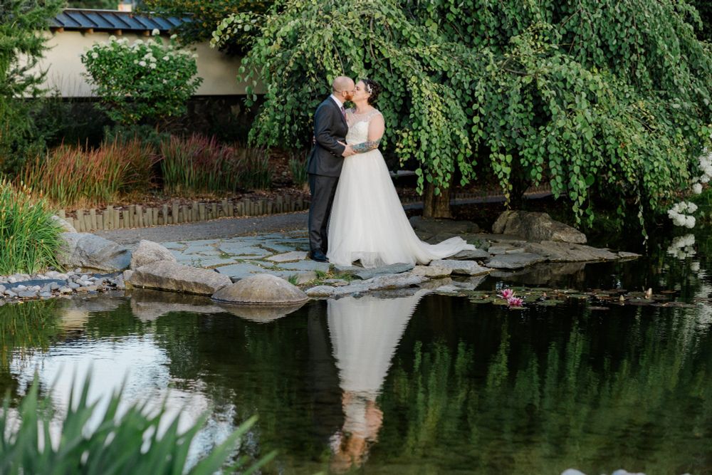 A bride and groom kissing in front of a lake with low trees around them. They are reflected very well in the water below