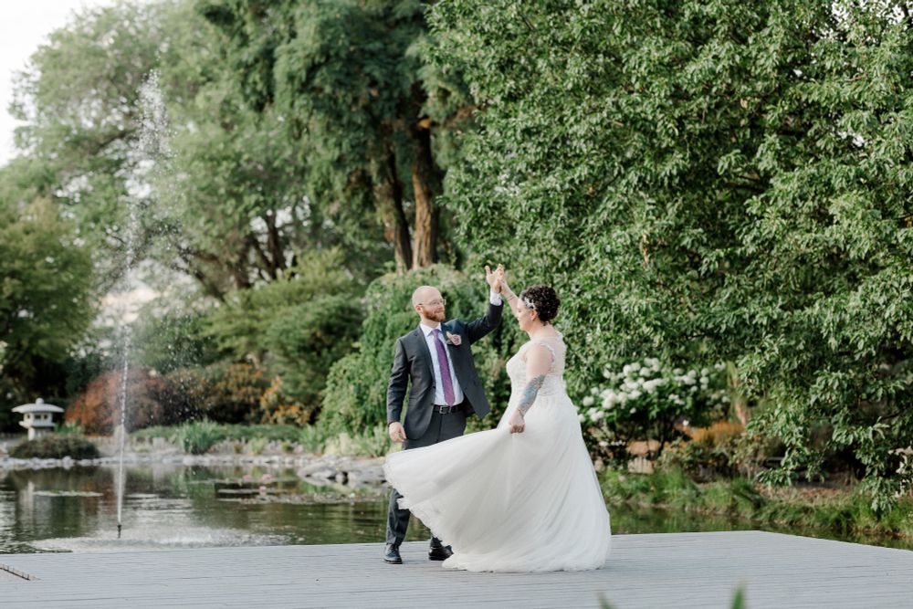 A bride and groom dancing on a dock in front of a pond with a fountain and surrounded by trees
