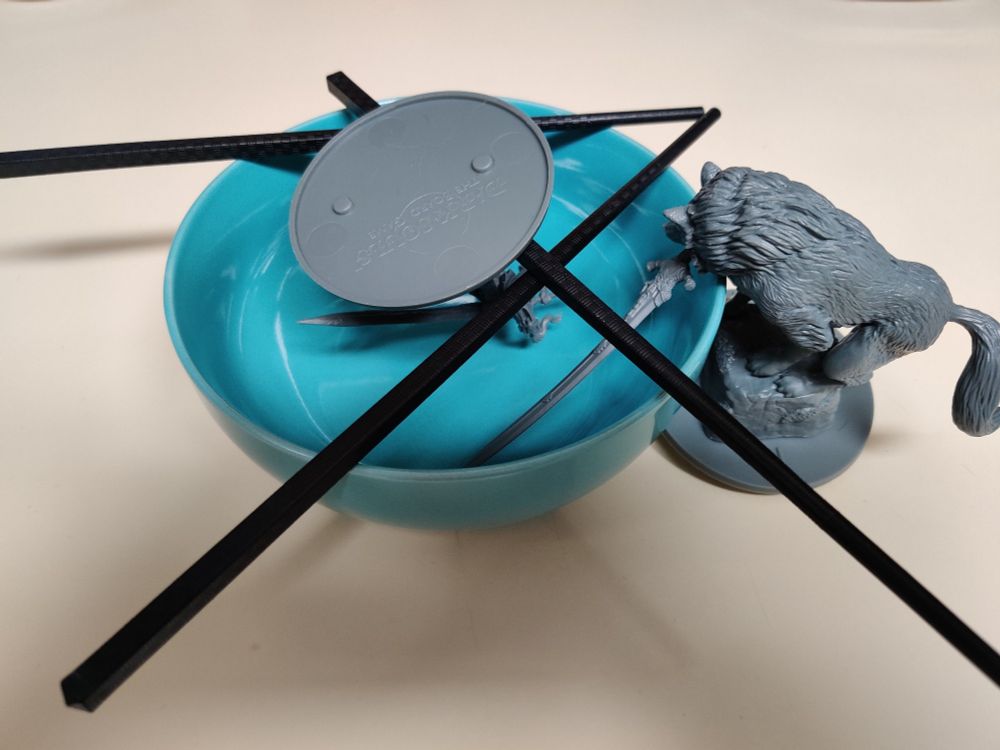 Sif and Artorias miniatures from the Dark Souls Board Game arranged around and in a bowl using chop sticks to suspend Artorias upside down. It's about to have near boiling water poured into it in the interest of straightening their swords.