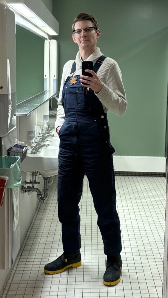 full-length selfie of me, a handsome man wearing navy blue overalls over a cream sweater and white polo, black boots with yellow soles, and a bigass brooch shaped like a maple leaf
