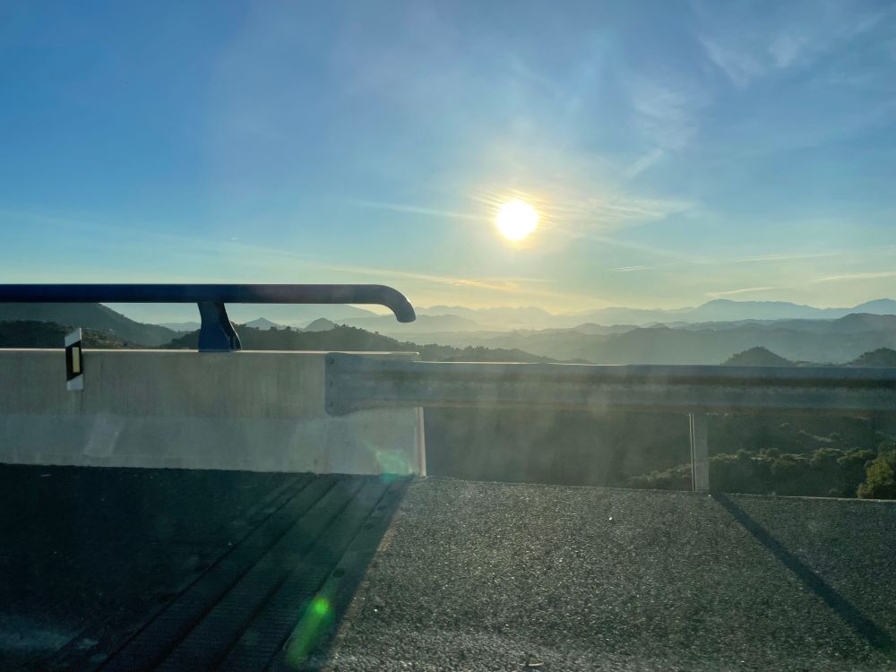 Tief stehende Sonne über Hügeln im Dunst. Zur Hälfte ragt von links eine Autobahnbrüstung ins Bild. 