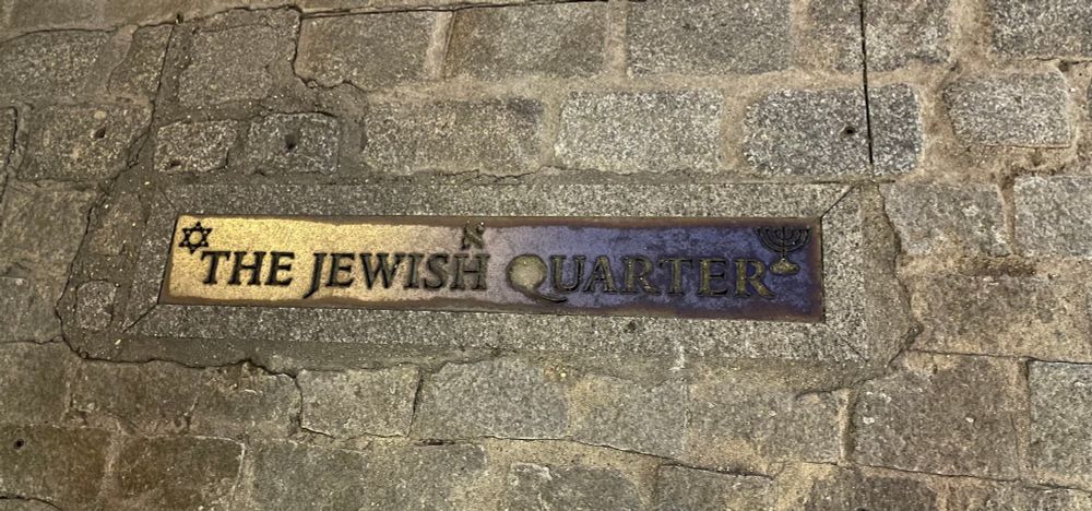 Eine Plakette im Kopfsteinpflaster erinnert an das jüdische Viertel. Text „The jewish quarter“ und ein Davidstern sowie ein Kerzenleuchter 