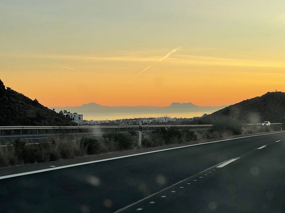 Blick durch die nach der langen Fahrt nicht mehr saubere Windschutzscheibe über die Autobahn und das Mittelmeer rüber nach Marokko unter dem orangefarbenen Abendhimmel. 