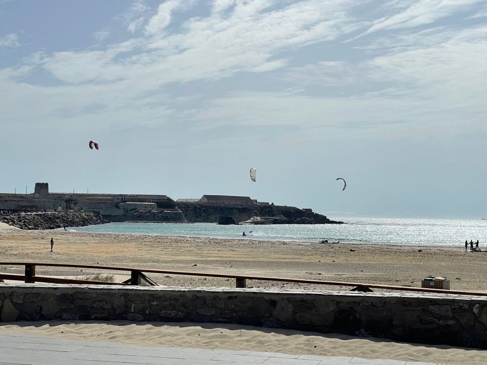 Blick von der Strandpromenade zur Isla de Palomas von Tarifa. Ein paar Kitesurfer sind unterwegs. Ihre Schirme stehen am bewölkten Himmel. 