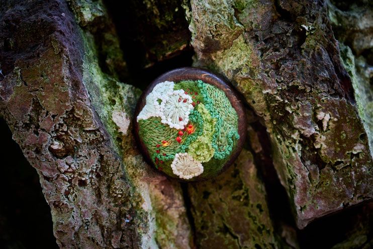 A small embroidered moss and lichen artwork rests against a textured rock background. The piece consists of embroideries in the shapes of white and green lichen plaques, felted moss, and scattered Trichia decipiens slime mold bulbs with embedded lights. The lights are colored red by the hue of the bulbs. The whole is mounted on a wooden ring.