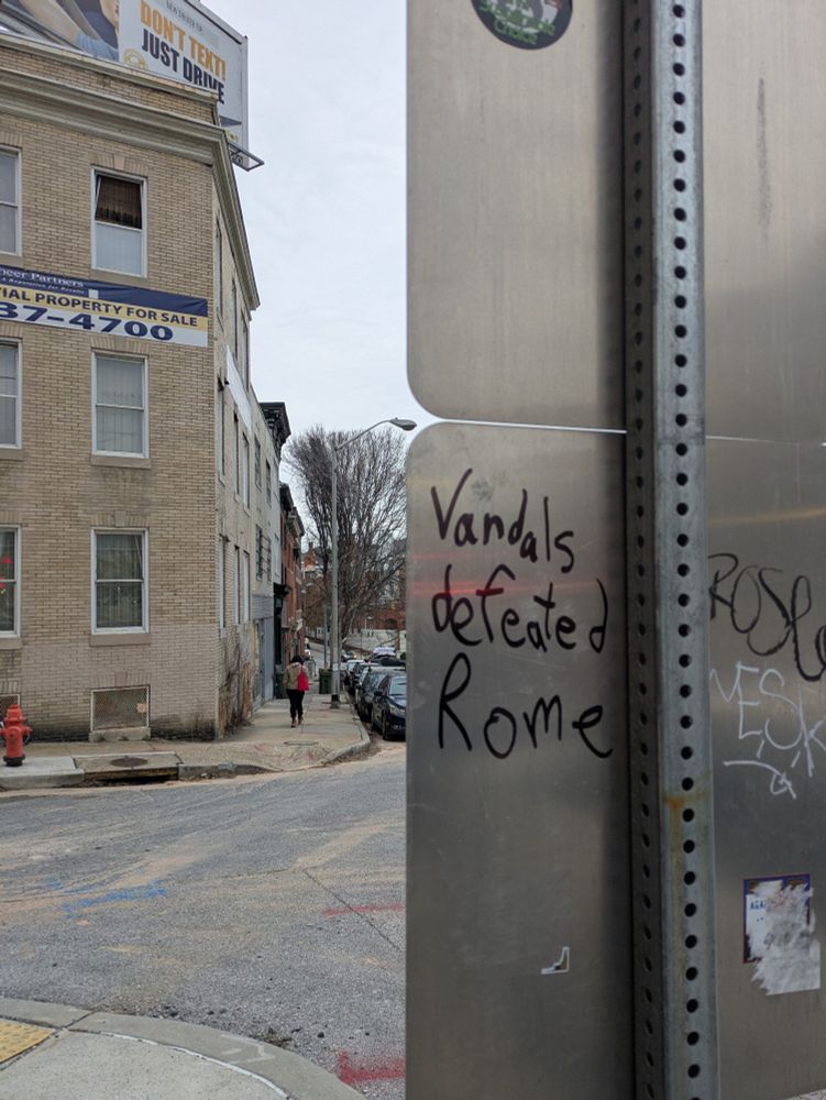The back of a metal sign is in the right side foreground with the phrase "Vandals defeated Rome" graffitied on it, vertically splitting the image. To left of the sign is the city streetscape beyond it, with a building, a person walking, parked cars and an orange fire hydrant visible.