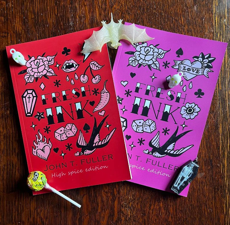 Two copies of Fresh Ink by John T Fuller show high spice version with red cover and low spice version with pink cover, both covers have black and white illustrations of tattoo flash. The books are surrounded by Halloween items like small plastic skulls, a rubber bat, and a lollipop