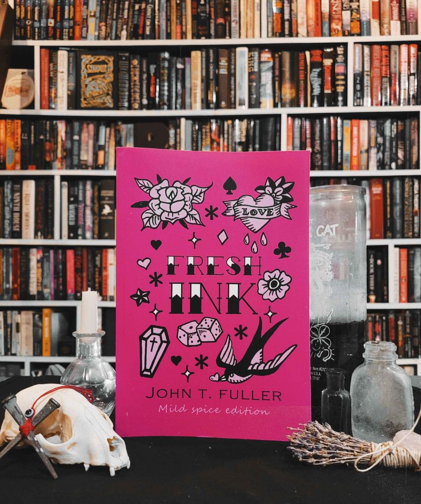 Fresh Ink mild spice has a pink cover with traditional flash tattoo designs in black, shown here surrounded by a cat skull, candles, herbs and in front of an impressive wall of book shelves
