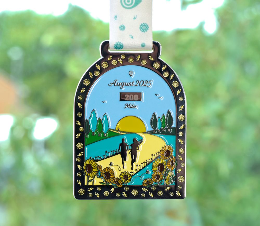 Image showing a metal medal (in the shape of an archway; hanging by a white and teal in colour ribbon) set against a blurred (green in colour) natural background. The medal features sunflowers, as well as two silhouettes of people running along a yellow road (set in nature with grass, trees, flowers, birds, a blue sky and a sunrise). The medal reads "August 2025"; "200 Miles".