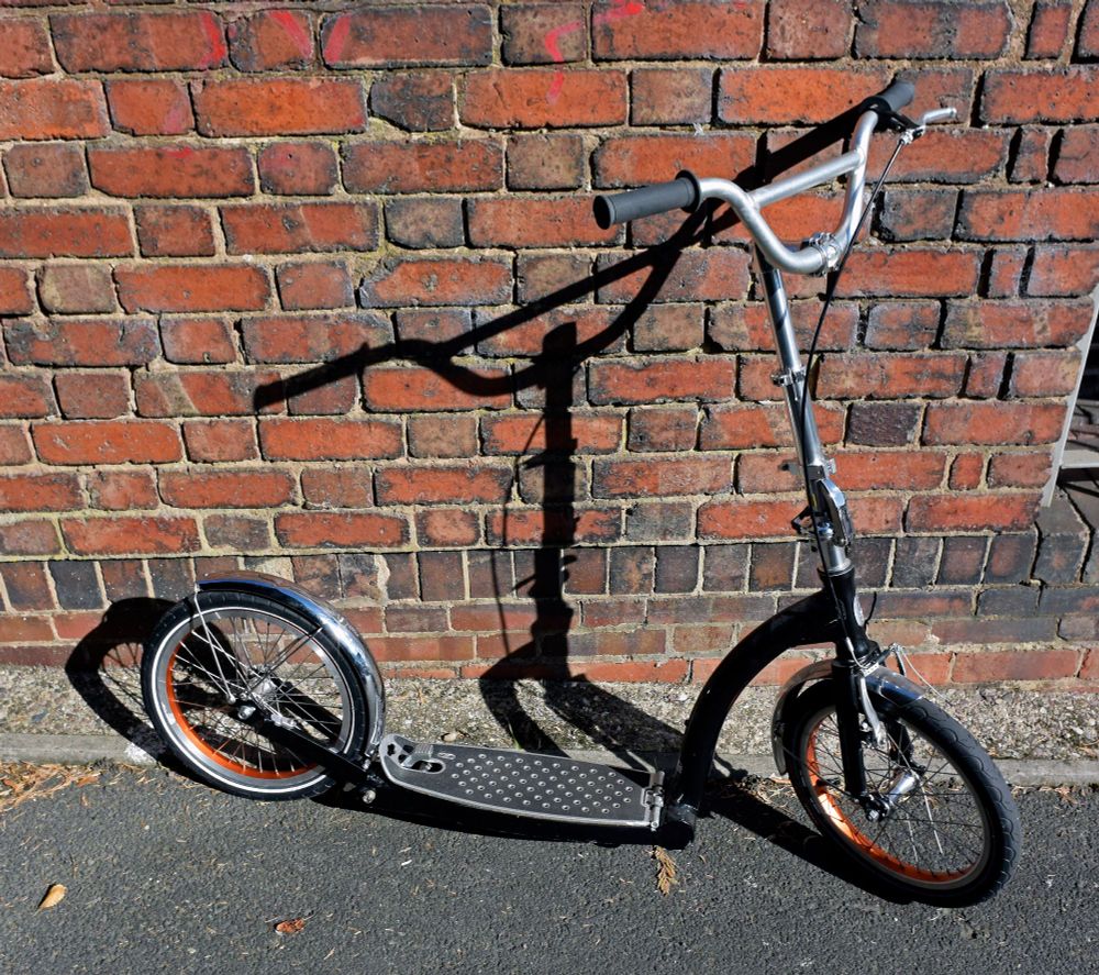 Image showing a folding adult kick scooter/kick bike that is black in colour with silver in colour applique (mudguards; stem; handlebar). The kick scooter is set against a red brick wall and sits upon a tarmac pavement.