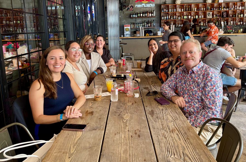 A group of smiling people pose for a photo. They are seated at a long wooden table. Behind them is a fully stocked bar. 