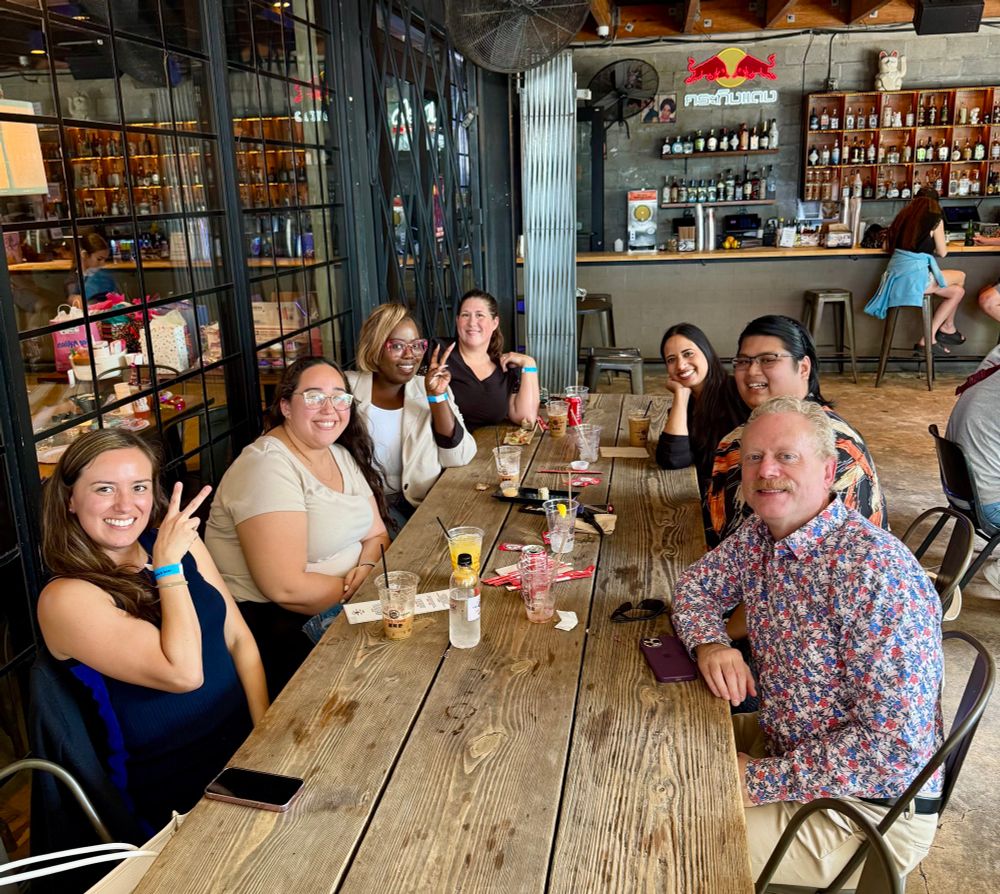 A group of smiling people pose for a photo. They are seated at a long wooden table. Behind them is a fully stocked bar. 