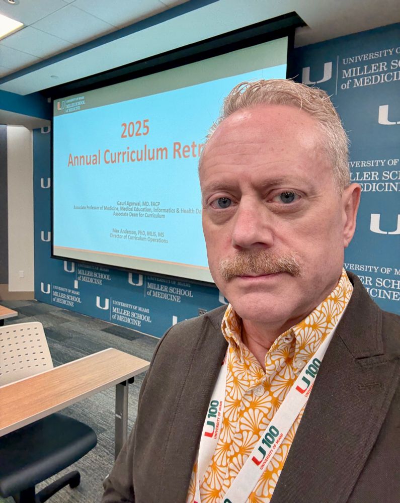 A man with deep under-eye bags is standing in front of a slide on a large screen. 