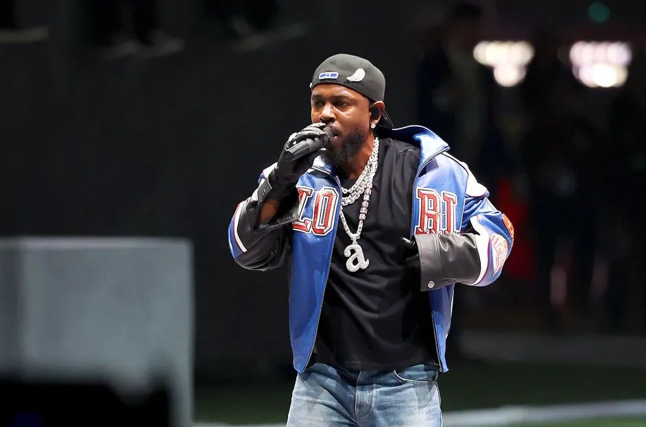 Kendrick Lamar performs at the Super Bowl LIX halftime show, wearing a backwards baseball cap, blue and black varsity-style jacket, and black shirt. Among his chain necklaces is a prominent serif letter 'a' pendant. He's speaking into a microphone with a focused expression, performing against a dark background with stage lighting.