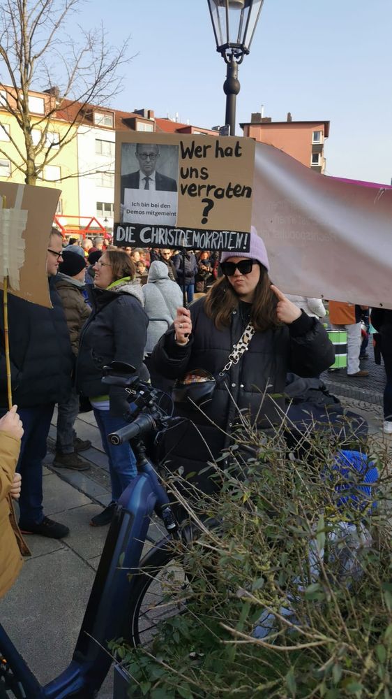 Ich stehe bei einer Demo gegen Rechts und halte ein Schild mit dem Text: Wer hat uns verraten? Die Christdemokraten! Auf dem Schild ist ein auf Papier ausgedrucktes Foto von Friedrich Merz. Er hält ein Schild in der Hand auf dem „Ich bin bei den Demos mitgemeint“ steht. 