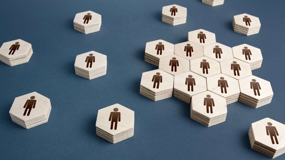 Stacks of wooden hexagons with human icons arranged on a dark surface, with one large clustered group symbolizing a team and several smaller, separated stacks representing individuals or smaller groups.