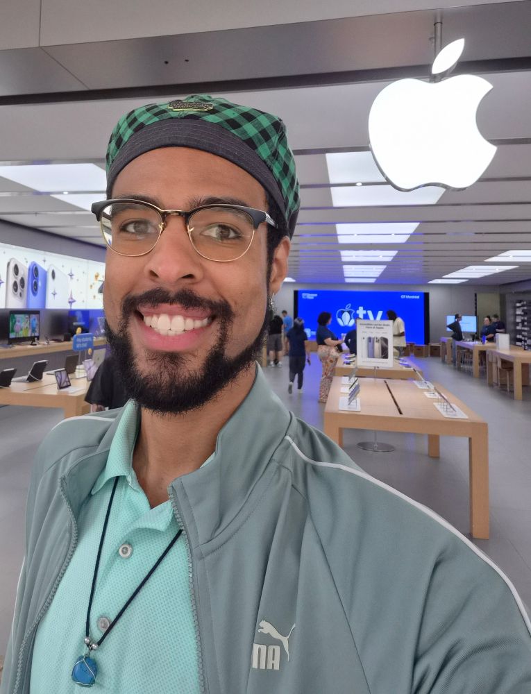 A smiling young Black adult male with glasses and facial hair wearing a green plaid Kangol hat, a sage green jacket, and a mint green polo shirt. He is also wearing a turquoise crystal on his necklace and standing in front of an Apple retail store.