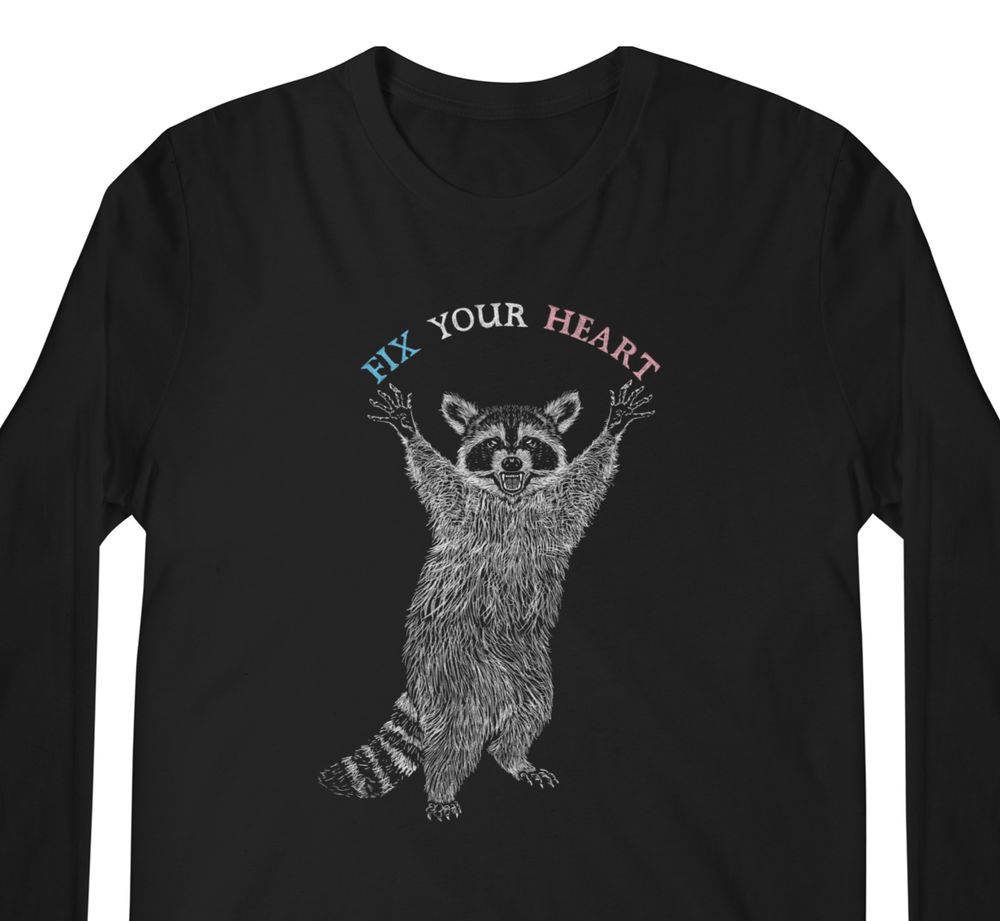 Photo of a long-sleeved black t-shirt with an illustration of a raccoon standing on its hind legs with front paws raised by its snarling face. The Words fix your heart in the blue, white, and pink colors of the trans flag are in a semi-circle between its paws.