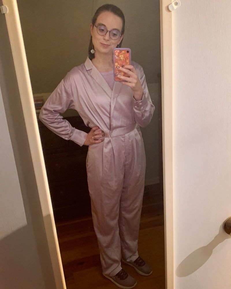 Kelsey is posing standing in front of a full length mirror that’s mounted on a white door. She’s wearing a satiny-fabric jumpsuit in lavender, disco ball dangling earrings, purple sneakers, and purple cat ear framed glasses.  