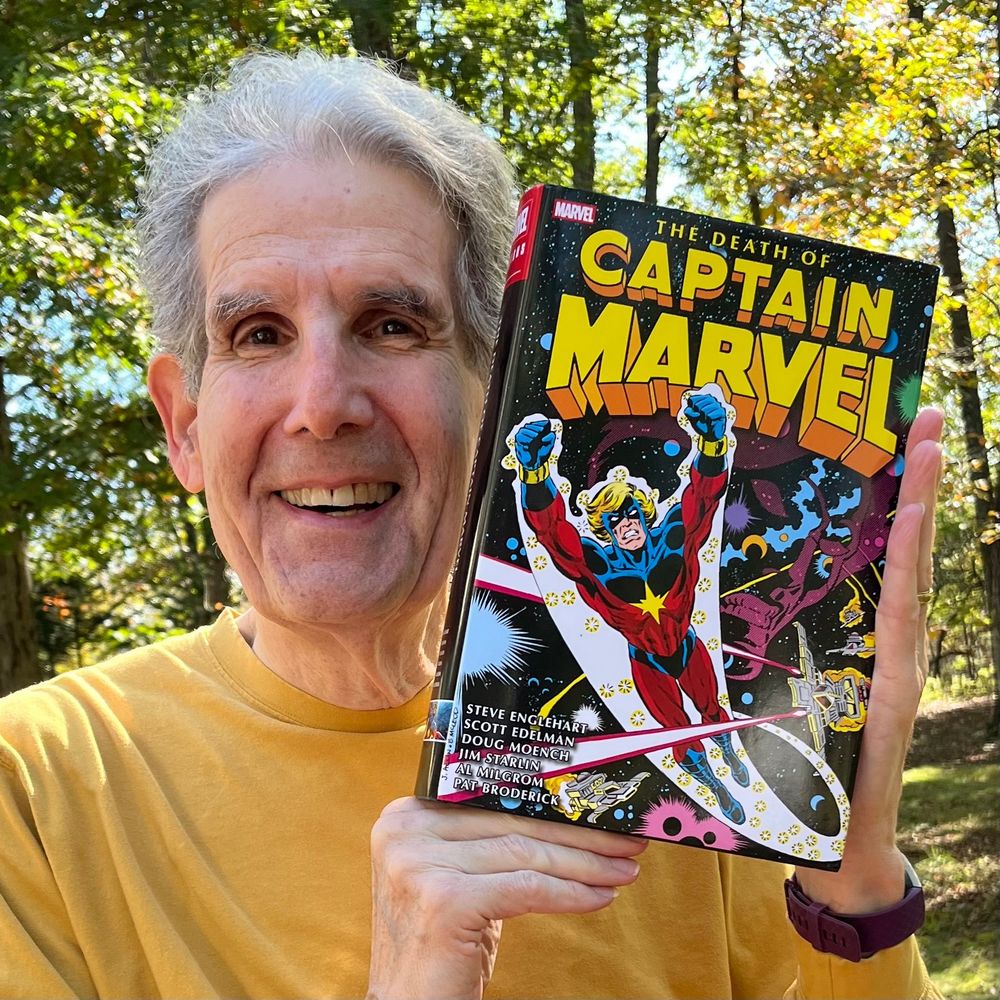 A photo of me in the backyard, trees behind me, holding my copy of the book near my face. I'm smiling and wearing a yellow, long-sleeved T-shirt. The cover shows Captain Marvel zooming around the spaceways, a trail of sparkles behind him. The names listed on the cover are:

Steve Englehart
Scott Edelman
Jim Starlin
Al Milgrom
Pat Broderick
