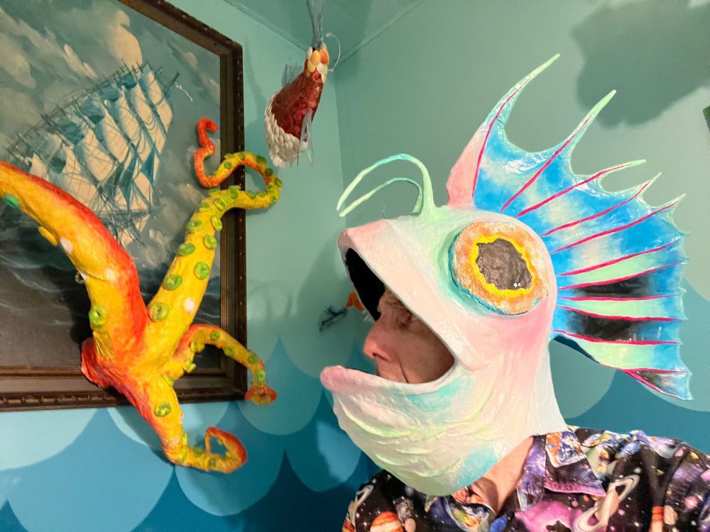 On the left, octopus tentacles of yellow and red with suckers of green reaching through the wall out of a painting go a sailing shop. On the right, me with my head encased on a fish's, only my eyes, nose, and upper lip visible. The head of the mask is opalescent, with pinks, and blues, and greens, and large yellow and red eyes with black pupils. There's a large tail fin coming out the back, with red veins. There are also two green antennae. The wall is painted with waves of various blues.