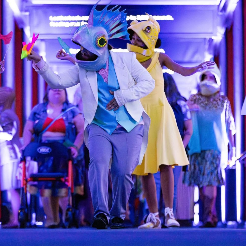 A person in what is now my fish head mask marching toward the camera. They are wearing a blue vest, pink shirt, and white jacket. The pants are perhaps lavender, but it's hard to say in the light of the aquarium. Though the mask cannot be seen in its full color due to that light, it's opalescent, with pinks, and blues, and greens, and large yellow and red eyes with black pupils. There's a large tail fin coming out the back, with red veins. There are also two green antennae. They's being followed by a person wearing a yellow dish mask and wearing a yellow dress.