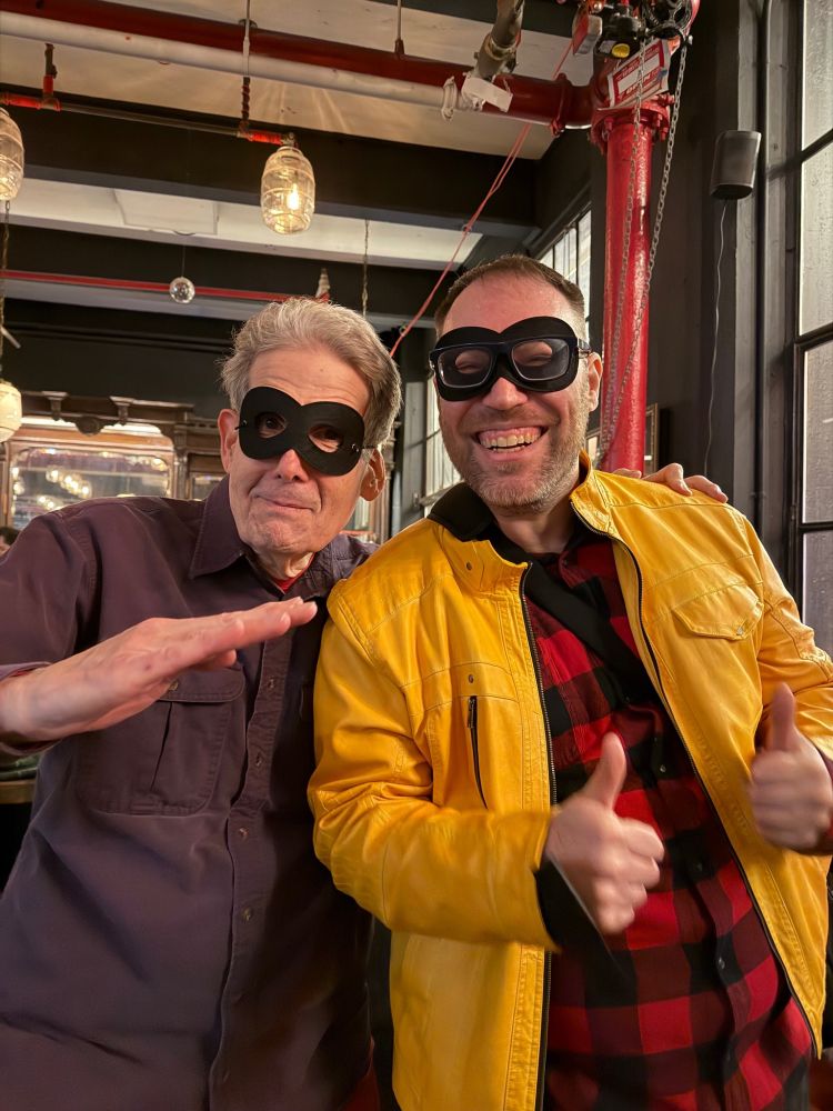 Two guys with stereotypical black burglar masks. I'm wearing a purple shirt and making a karate chop with my right hand, while Michael is wearing a yellow jacket over a red plaid shirt and giving two thumbs up. Michael's mask is under his glasses.