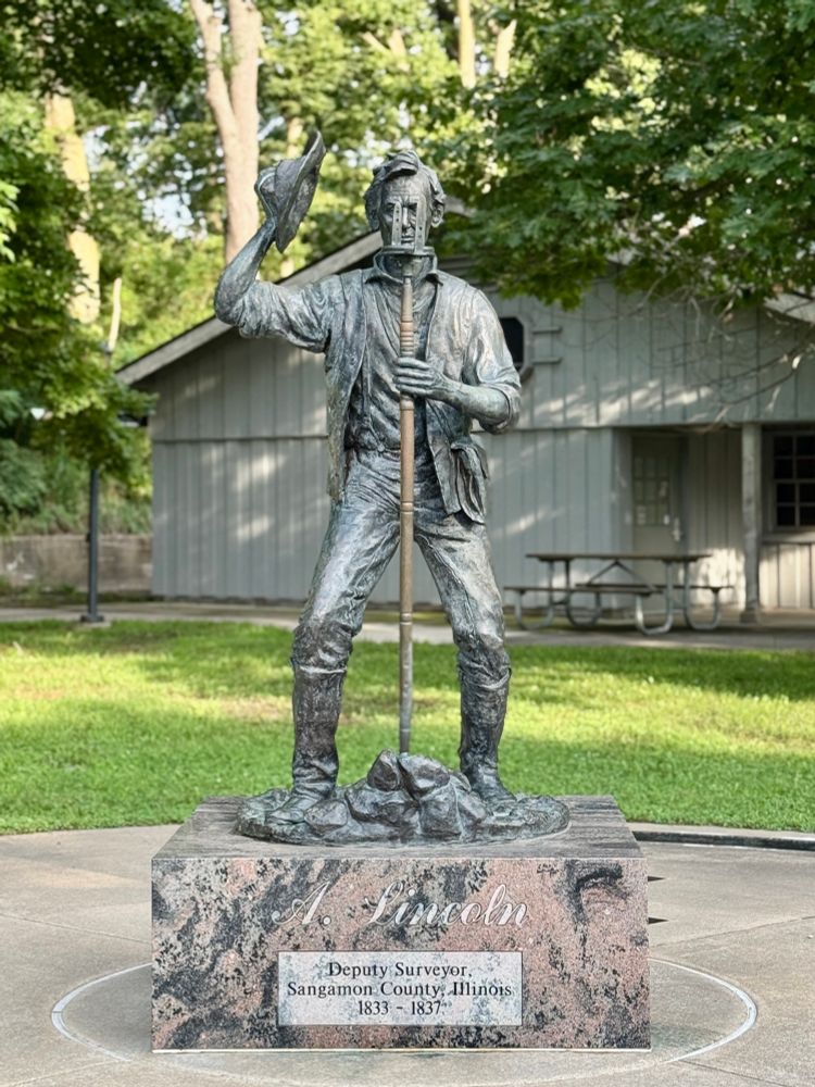 Statue of Lincoln brandishing a short, wide-brimmed cap, bending his knees a bit, and peering at what I think is some sort of surveyor’s compass