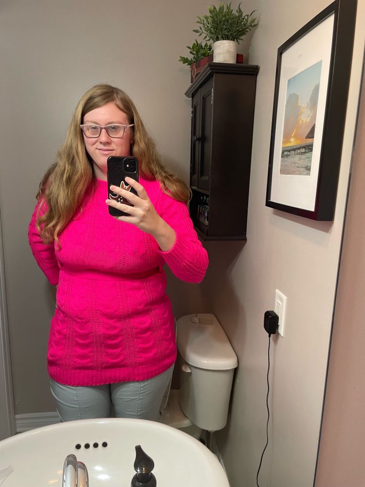 An image of a woman with long, dark blonde hair wearing a bright pink sweater with long sleeves. She is taking the photo in a bathroom mirror so she is holding her phone in the image.